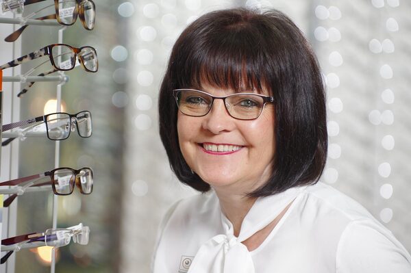 Freundliche Optikerin mit Brille steht neben einer Auswahl moderner Brillengestelle.